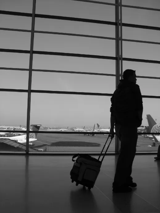 A packed rolling suitcase beside an airport terminal window at trip departure, illustrating a traveler weighing using a travel agent versus booking online with Suncoast Travel in Plymouth, Michigan.